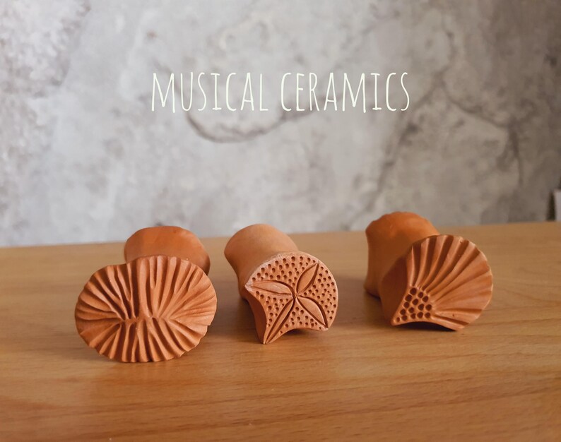 May include: Three terracotta-coloured ceramic stamps with distinct carved patterns on their bases. The left stamp features a radial line design, the centre stamp has a four-petal floral motif, and the right stamp shows a fan-like pattern. Text reads MUSICAL CERAMICS.