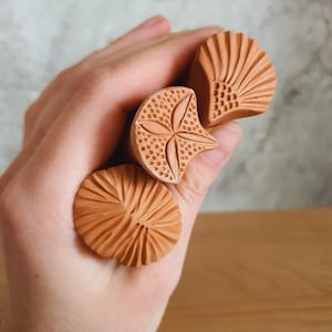 May include: Three terracotta-coloured clay stamps with distinct carved patterns, including a fan shape, a star-like design with dotted textures, and a radial line pattern. The text MUSICAL CERAMICS is visible at the top of the image.