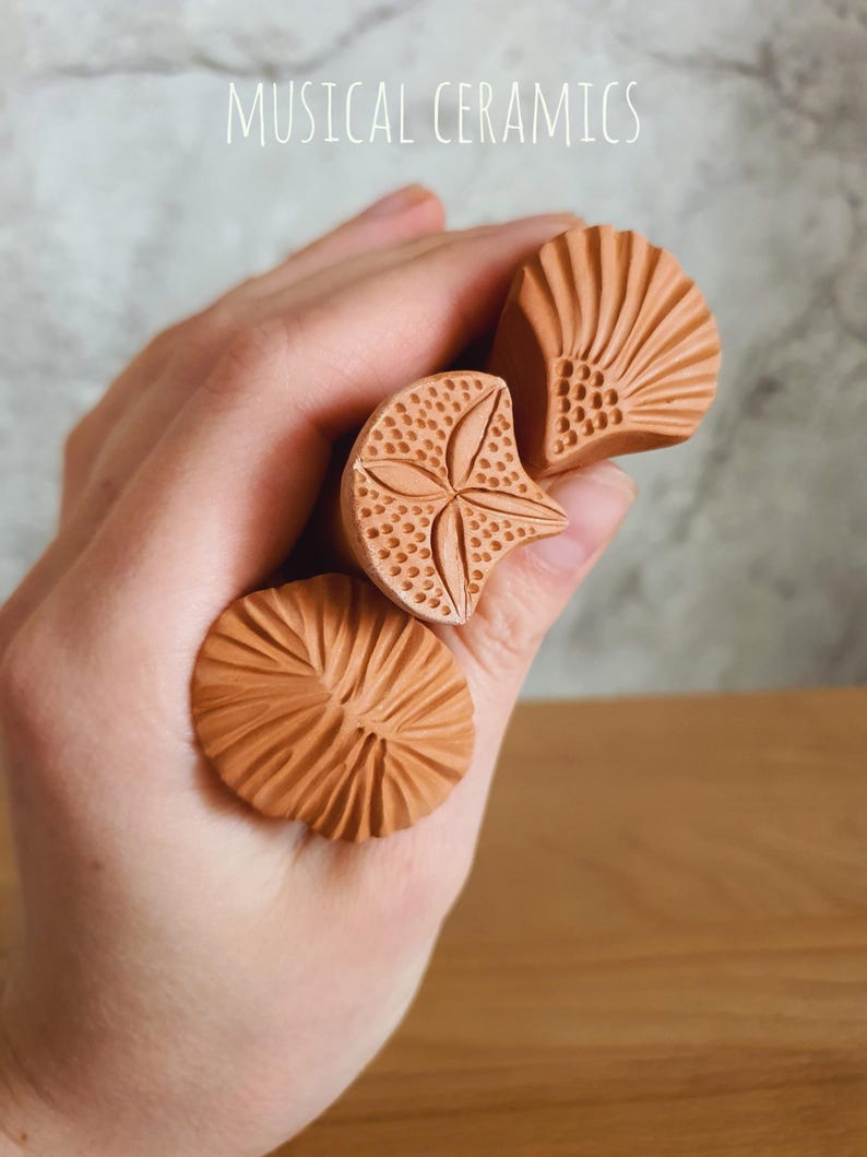 May include: Three terracotta-coloured clay stamps with distinct carved patterns, including a fan shape, a star-like design with dotted textures, and a radial line pattern. The text MUSICAL CERAMICS is visible at the top of the image.