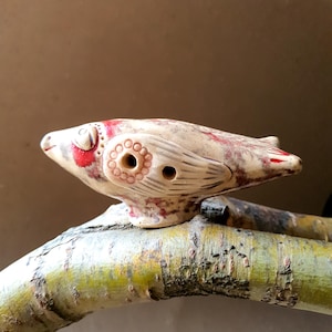 Puede incluir: Un silbato de cerámica con forma de pájaro pintado en rojo y blanco, con un agujero para silbar. El pájaro está posado en una rama.
