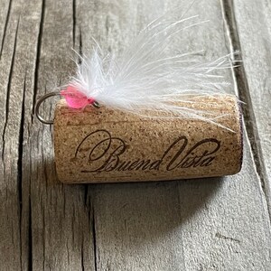 May include: A white feather fly fishing lure with a pink bead tied to a wine cork with the words "Buena Vista" engraved on it.