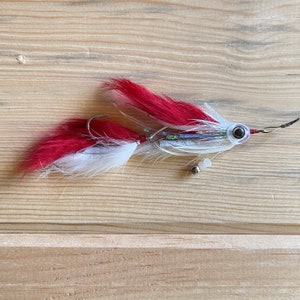 May include: A red and white fly fishing lure with a holographic finish and a bead chain. The lure has a single hook and a black eye.