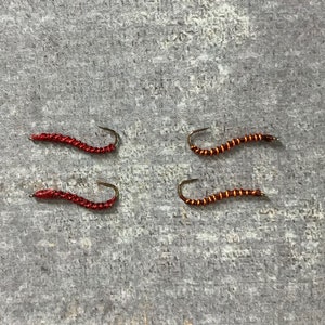 Puede incluir: Cuatro señuelos de pesca con mosca, dos rojos y dos naranjas, con un anzuelo en cada extremo.
