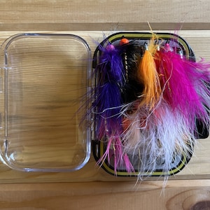 May include: A clear plastic box containing a selection of colourful fly fishing lures. The lures are made with feathers and have different coloured beads and bodies.