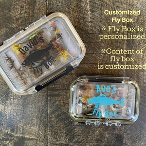Może przedstawiać: Dwie przezroczyste plastikowe pudełka na muchy z personalizowanymi etykietami. Górne pudełko ma czarną sylwetkę ryby i napis "David's Fly Box". Dolne pudełko ma niebieską sylwetkę ryby i napis "Ava's Fly Box".