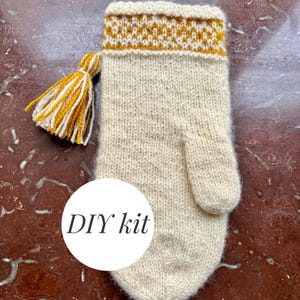 Pode incluir: Uma luva de malha creme com um borla amarela e branca e um punho estampado. A luva tem um polegar pequeno e as palavras "DIY kit" num círculo branco.