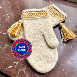 May include: A pair of cream-colored knitted mittens with a decorative band of yellow and brown at the wrist. Each mitten has a tassel of yellow and white yarn. The mittens have the text "SÁMI MADE" in a circular logo.