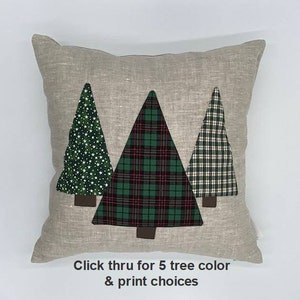 Puede incluir: Una almohada de lino beige con tres árboles de Navidad aplicados. Los árboles están en diferentes patrones: verde con estrellas blancas, verde a cuadros y vichy verde y blanco. El texto "Haga clic para ver 5 colores y diseños de árboles" está en la parte inferior de la almohada.