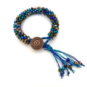May include: A beaded bracelet with a copper-colored button clasp and a tassel. The bracelet is made of small, colorful beads in shades of blue, green, and gold. The tassel is made of blue threads with decorative beads.
