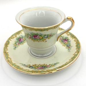May include: A white teacup and saucer set with a floral pattern in shades of pink, green, and yellow. The set has a gold trim around the edges.
