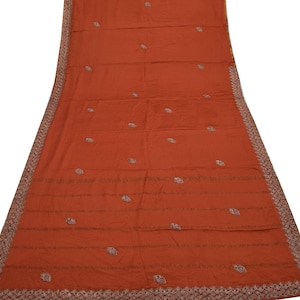 Peut inclure: Un textile orange brûlé avec des détails brodés. Le tissu présente de petits motifs floraux dispersés et des lignes horizontales. La bordure est finement conçue avec un motif géométrique répétitif dans des tons de marron et de crème. Le textile est étalé, montrant toute sa longueur.