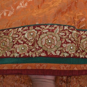 Puede incluir: Primer plano de una sección de tela con un borde floral adornado. El borde presenta flores doradas bordadas con detalles de lentejuelas sobre un fondo burdeos, con rayas verdes y naranjas. El fondo es una tela naranja texturizada.