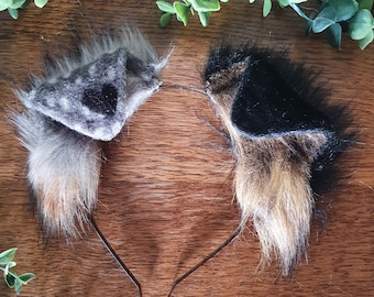 May include: A pair of furry cat ears headband with a black and grey ear on the left and a brown and black ear on the right. The headband is black.
