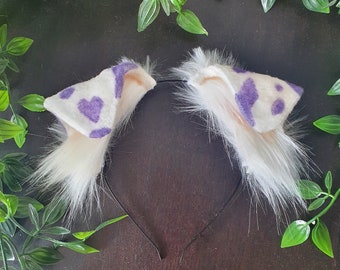 May include: A pair of white furry cat ears with purple and white spots on the ears. The ears are attached to a black headband.