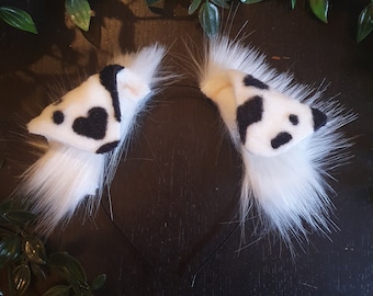 May include: A pair of white furry cat ears with a black and white cow print fabric. The ears are attached to a black headband.