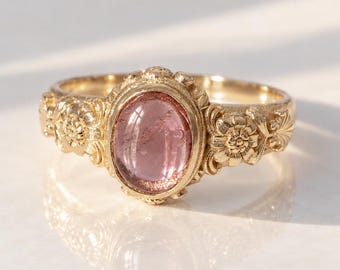 May include: A gold ring featuring a central oval-shaped pink gemstone set in a bezel. The band is decorated with intricate floral carvings on both sides of the setting, showcasing a polished metallic finish and a smooth, rounded inner band.