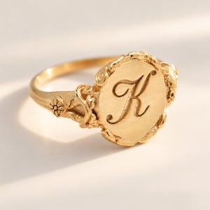 Op de afbeelding: Een gouden ring met een rond plaatje waarop de letter K in cursief schrift is gegraveerd. De ringband en de zetting van het ronde plaatje zijn versierd met gedetailleerde, verhoogde bloem- en krulmotieven in een matte goudkleur.