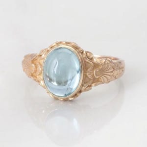 Op de afbeelding: Een goudkleurige ring met een ovale, lichtblauwe edelsteen. De ring heeft een sierlijk ontwerp met bloemen- en schelpachtige patronen. De edelsteen is gezet in een gouden bezel en de band heeft een getextureerde afwerking.