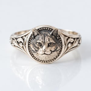 May include: A silver-toned metal ring featuring a circular centerpiece with a detailed, raised relief of a cat face. The circular frame is bordered by small beads, and the band is decorated with intricate, symmetrical leaf-like engravings.