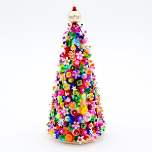 Könnte beinhalten: Ein konischer dekorativer Baum, der mit einer dichten Anordnung aus bunten Perlen, Pailletten und Kunststoffblumen in den Farben Pink, Orange, Gelb, Grün, Blau und Lila bedeckt ist und von einer goldenen Kugel sowie einer kleinen roten Perle gekrönt wird.