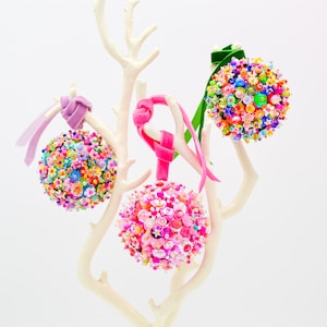 May include: Three spherical ornaments covered in colorful beads, sequins, and small floral embellishments. Each ornament hangs from a velvet ribbon in lavender, hot pink, or green, suspended from a white, branch-like display stand.