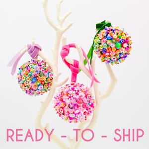 Peut inclure: Trois ornements sphériques recouverts de perles colorées, de paillettes et de petites breloques florales, suspendus par des rubans de velours sur un support blanc en forme de branche. Le texte READY - TO - SHIP est imprimé en rose.