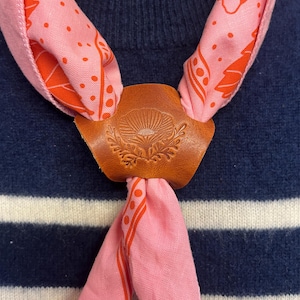 Könnte beinhalten: Ein rosa Schal mit orangefarbenem Muster wird von einem braunen Lederverschluss gehalten. Der Verschluss hat ein geprägtes Blumendesign. Der Schal wird über einem marineblau-weiß gestreiften Pullover getragen.