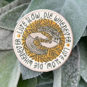 May include: A white embroidered patch with green and yellow foliage and two stylized figures. The text reads "Live Slow, Die Whenever You Die, Live Slow".