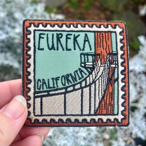 May include: Embroidered patch featuring a bridge scene with the text "EUREKA CALIFORNIA" in black. The patch has a postage stamp design with a light green background and a brown border. The patch is square and has a textured appearance.