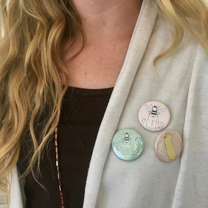 May include: Three round pins with bee designs. The top pin is pink with a bee and the text "Bee Kind". The middle pin is green with a bee and flowers. The bottom pin is yellow with a bee and the outline of California.