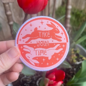 May include: A pink and orange round embroidered patch with two white rabbits running in opposite directions. The patch says "Take your time" with stars around the rabbits.