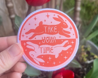 Könnte beinhalten: Ein runder gestickter Patch in Rosa und Orange mit zwei weißen Kaninchen, die in entgegengesetzte Richtungen laufen. Der Patch sagt "Nimm dir Zeit" mit Sternen um die Kaninchen herum.