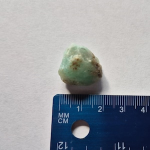 May include: A small, rough-cut, light green gemstone with brown inclusions. The stone is positioned next to a blue ruler, with measurements in millimeters and centimeters.