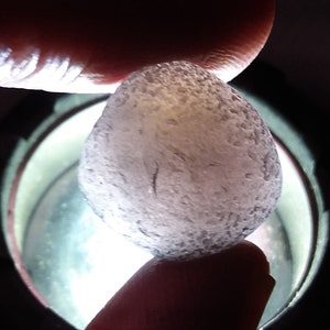 May include: A close-up of a smooth, white, round stone with a slightly translucent appearance. The stone is being held by a finger and is illuminated by a bright light.
