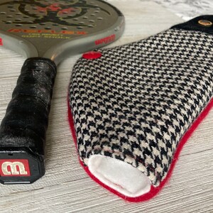 May include: A black and white patterned racquet cover with a red trim and a black top. The cover has a small black button on the top. The cover is lying on a wooden surface next to a silver racquet with a black handle.