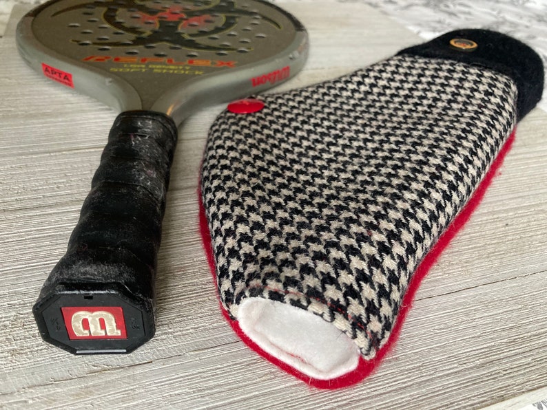 May include: A black and white patterned racquet cover with a red trim and a black top. The cover has a small black button on the top. The cover is lying on a wooden surface next to a silver racquet with a black handle.