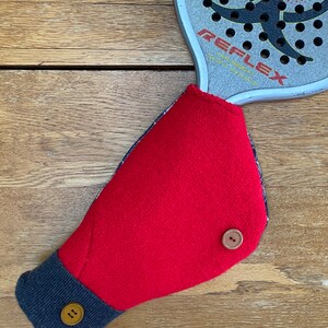 May include: Red fleece hand warmer with a navy blue cuff and two brown buttons. The hand warmer is designed to fit over a racquet handle.