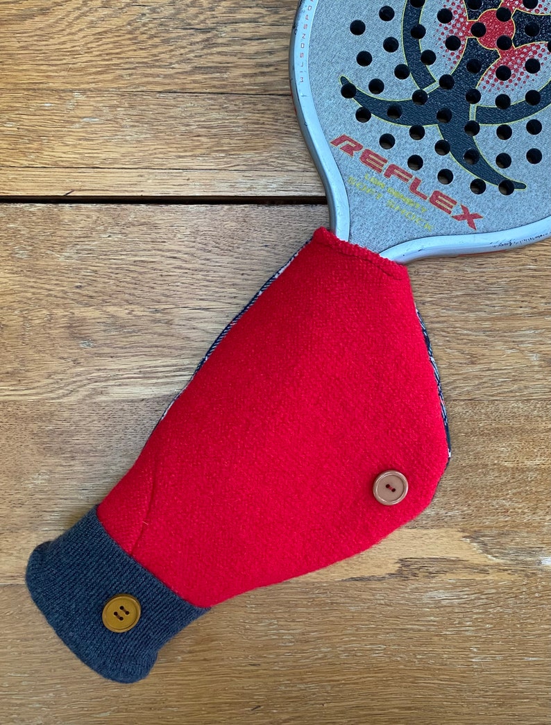 May include: Red fleece hand warmer with a navy blue cuff and two brown buttons. The hand warmer is designed to fit over a racquet handle.