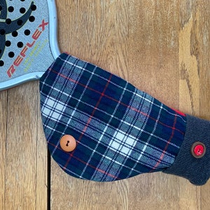 May include: A blue, white, and red plaid fabric arm warmer with a brown button and a gray cuff. The arm warmer is on a wooden surface.