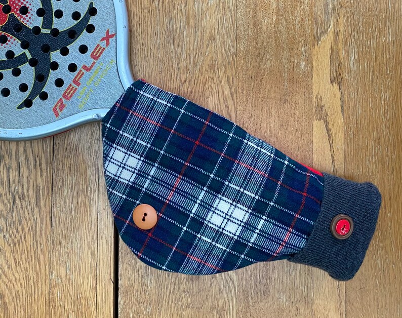May include: A blue, white, and red plaid fabric arm warmer with a brown button and a gray cuff. The arm warmer is on a wooden surface.