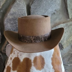 May include: A tan felt hat with a flat top and a wide, curved brim. The hat features a dark brown band around the base with a textured, cracked pattern that resembles leather. The hat is positioned atop a white and brown cowhide patterned surface.