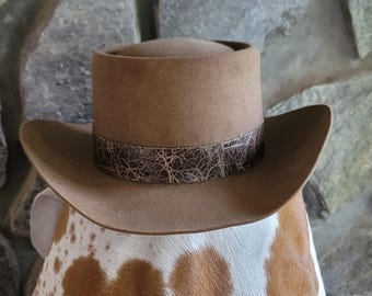 May include: A tan felt hat with a flat top and a wide, curved brim. The hat features a dark brown band around the base with a textured, cracked pattern that resembles leather. The hat is positioned atop a white and brown cowhide patterned surface.