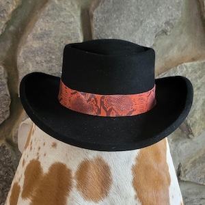 May include: A black hat with a wide brim and a snakeskin-patterned band in shades of orange and red. The hat is set against a stone background. The hat's design is a classic style.