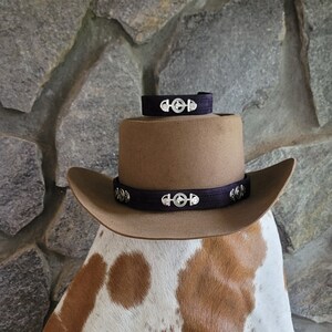 May include: A brown felt cowboy hat with a black leather band and silver conchos. The hat is sitting on a white and brown cowhide.