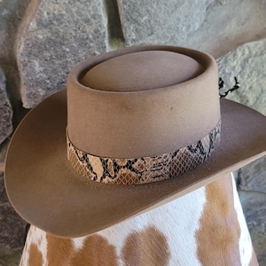 May include: A tan cowboy hat with a snakeskin band. The hat has a rounded crown and a wide brim. The snakeskin band features a pattern of brown, black, and tan. The hat is set against a stone background.
