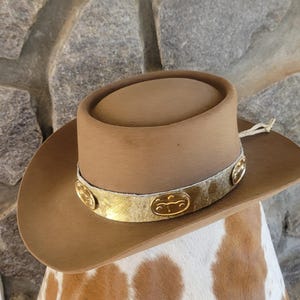 May include: A brown cowboy hat with a gold band featuring decorative accents. The hat has a rounded crown and a wide brim. The hat is displayed on a cowhide surface.