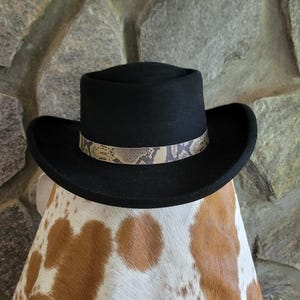 May include: A black fedora hat featuring a flat brim and a structured crown. The hat is accented with a decorative band that displays a tan and dark brown snakeskin pattern, which wraps around the base of the crown above the brim.