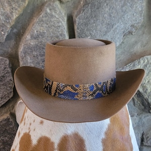 May include: A brown cowboy hat with a snakeskin band featuring blue and black accents. The hat has a wide brim and a rounded crown. The hat is resting on a spotted surface.