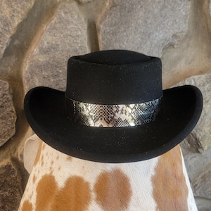 May include: A black hat with a snakeskin patterned band. The hat has a flat top and a wide brim. The snakeskin band is silver and gold. The hat is likely made of felt or a similar material.