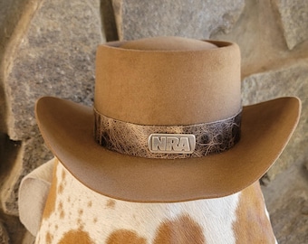 May include: A tan cowboy hat with a brown leather band featuring the letters "NRA" in a silver emblem. The hat has a wide brim and a rounded crown. The hat is resting on a cowhide pattern.